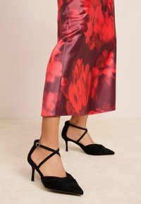 Mustad seemisnahast kõrgete kontsadega kingad terava varba, esiosas sõlmitud detaili ja pahkluu rihmadega. Kandmiseks koos punase lillelise satiinse seelikuga.