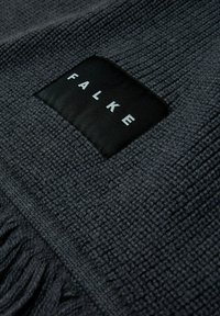 Tissu tissé sombre avec une surface texturée. Présente une étiquette rectangulaire noire avec des lettres blanches indiquant "FALKE". Bords frangés.
