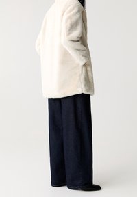 Crème faux bont jas met een relaxed fit, voorzien van zijzakken, gecombineerd met donkerblauwe wijde jeans en zwarte schoenen.