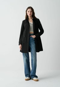 Černý trenčkot s dvouřadým designem, detaily s knoflíky a uvolněným střihem. Vybavený pruhovaným crop topem a rozšířenými džíny.