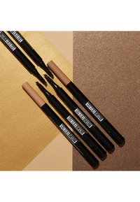 Quatre feutres à sourcils avec des corps noirs et des bouchons beige, dotés de pointes fines pour une application précise, disposés sur un fond doré texturé.