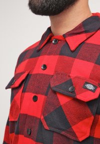 Camisa de franela a cuadros rojos y negros con frente abotonado, cuello puntiagudo y dos bolsillos en el pecho con botones negros.