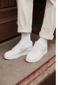 Person som bär vita Nike-sneakers med beige swoosh, vita ribbade strumpor och uppvikta gräddfärgade byxor står på en betongtrappa.