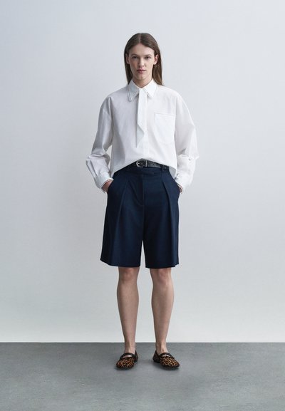 Femme debout contre un mur uni portant une chemise blanche à manches longues, un short marine à hauteur des genoux, et des mocassins à imprimé léopard, les mains dans les poches.