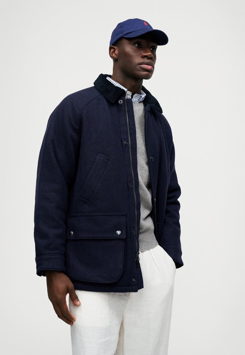 Veste en mélange de laine bleu marine avec un col en velours côtelé, des poches avant et des boutons-pression. Portée sur un pull gris et un pantalon blanc.