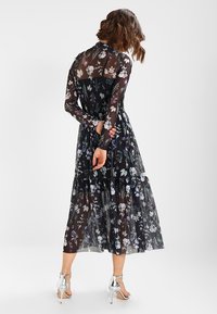 Robe noire à fleurs avec de longues manches transparentes, design en étages et motifs floraux contrastés en violet et blanc. Portée avec des talons argentés.