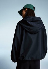 Zwarte hoodie met een relaxed fit, met een matte textuur. Een groene pet maakt de look compleet, gezien vanaf de achterkant tegen een lichte achtergrond.