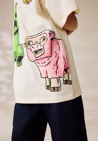 Baumwoll-T-Shirt in Cremefarbe mit lebendigen rosa und grünen Tiergrafiken; zeigt ein Schaf und eine Kreatur, betont kräftige Farben und Texturen.