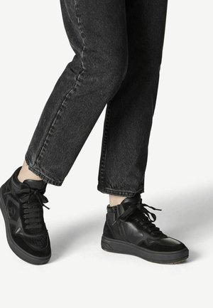 Osoba nosząca czarne wysokie trampki i czarne dżinsy na jasnym tle, pokazująca stopy i dolne partie nóg.