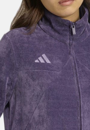 Chaqueta de forro polar en un material texturizado de color púrpura con cuello alto y cierre de cremallera. Presenta el logo de Adidas en un púrpura más claro en la parte superior izquierda.
