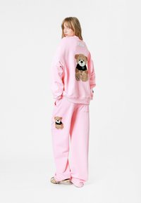 Conjunto de sudadera y pantalones de chándal rosa con apliques de oso de peluche en marrón y negro en la parte posterior y en la pierna del pantalón. Textura de tela suave.