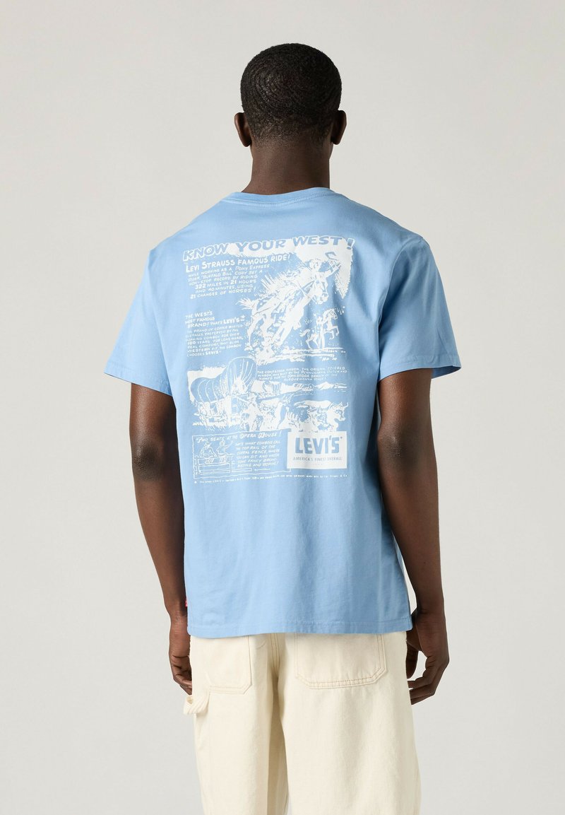 Mann trägt hellblaues T-Shirt mit weißer Vintage-Western-Grafik von Levi's auf dem Rücken, kombiniert mit hellbeigen Shorts.
