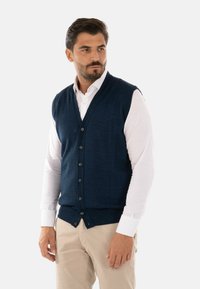 Gilet lavorato a maglia blu navy con scollatura a V, dotato di sei bottoni e orlo a costine. Indossato sopra una camicia bianca a maniche lunghe, abbinato a pantaloni beige.