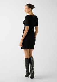 Schwarzes, geripptes Strickkleid mit kurzen Ärmeln und figurbetonter Silhouette. Bemerkenswerte Details sind ein dezentes Logo auf der Rückseite und ein ausgestellter Saum.