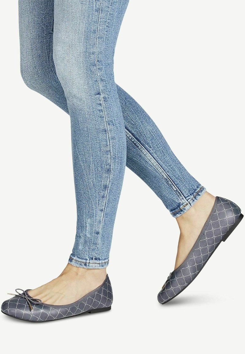 Pieds portant des ballerines gris matelassées avec de petits nœuds, associées à un jean skinny bleu clair sur fond uni.