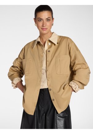 Frau mit zurückgebundenen Haaren, trägt eine beige Jacke mit Knöpfen über einer Spitzenbluse und einem schwarzen Lederrock, Hände in den Jackentaschen, neutraler Hintergrund.