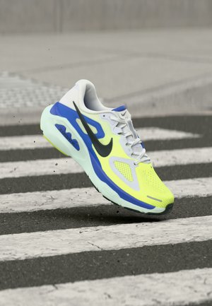 Chaussure de course Nike jaune vif, bleue et blanche en suspension au-dessus d'un passage pour piétons sur l'asphalte, avec de petits débris soulevés en dessous.