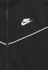 Černá mikina na zip z hladkého materiálu. Obsahuje bílé logo Nike a reflexivní designový prvek na hrudi.