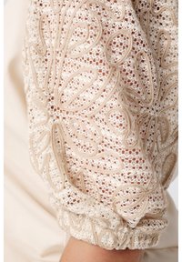 Kanten blouse met sleevem met beige borduurwerk op een lichtcrème achtergrond, voorzien van intrigerende bloemenpatronen en een geraffineerde manchet.