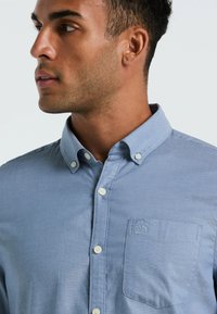 Helles blaues Hemd mit Knöpfen verfügt über einen Kragen, eine Brusttasche mit Logo und ein dezentes strukturiertes Muster. Die Knöpfe sind weiß und gleichmäßig verteilt.