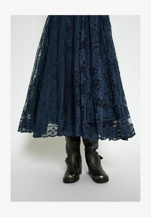 Tummansininen pitsinen midihame, jossa on monimutkaisia kuvioita ja aaltoileva helma, yhdistettynä mustiin nahkaisiin nilkkureihin, joissa on hihnat yksityiskohtina.