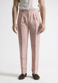 Pantalones de lino rosa claro con pliegues frontales, cierre de botón y bolsillos laterales. Combinados con zapatos marrones de tipo slip-on. Diseño minimalista.