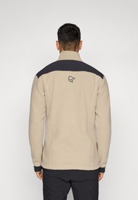 Beige fleecejakke med høy krage, mørkeblå skuldererme og en liten logo på baksiden. Myk tekstur og vanlig passform.