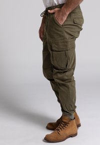 Olivgrüne Cargo-Hosen mit elastischem Kordelzugbund, schmal zulaufenden Beinen, mehreren Taschen und einem weichen, strukturierten Stoff. Kombiniert mit braunen Stiefeln.