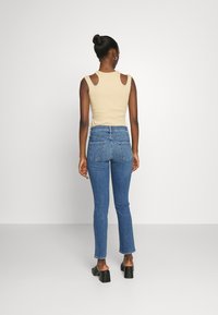 Top sin mangas de color beige con detalles recortados en los hombros, combinado con jeans azules de cintura alta y sandalias negras de tacón cuadrado. Vista posterior.
