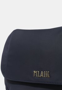Borsa blu navy con una superficie liscia, caratterizzata da un logo dorato e una chiusura a patta. Gli accenti cuciti lungo i bordi arricchiscono il design.