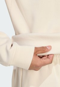 Krämfärgad sweatshirt med mjuk textur, ribbade muddar och avslappnad passform, med en hand som greppar midjebandet.