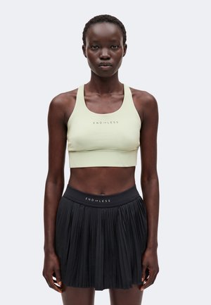 Vrouw draagt een lichtgroene sportbeha en een zwarte geplooide rok, beide met tekst "ENDLESS", staand tegen een effen lichte achtergrond.