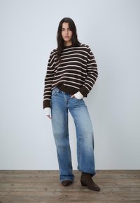 Brązowy sweter w paski z szerokim okrągłym dekoltem, w połączeniu z jasnoniebieskimi rozszerzanymi dżinsami i ciemnymi zamszowymi botkami. Tło z drewnianą podłogą.