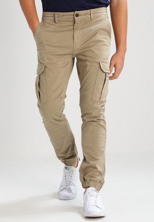 Cargo pants in beige, made of lightweight fabric. Features multiple side pockets, elastic cuffs, and a fitted waist. Worn with white sneakers.