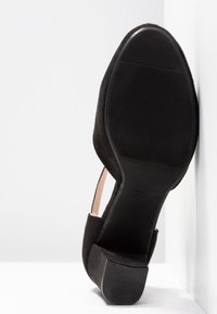 Chaussure femme en suede noir avec talon bloc, vue depuis le côté de la semelle, reposant contre un mur et un sol blancs.