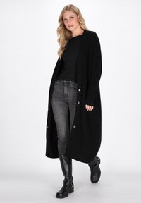 Cardigan lungo nero in tessuto a coste, con grandi bottoni, indossato sopra una maglietta nera e jeans grigi, abbinato a stivali neri fino al ginocchio.