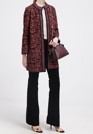 Vrouw in een lang jasje met een rood en zwart patroon, zwarte broek, goudkleurige open teen hakken, en een donkerrode leren handtas.