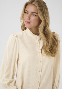 Ecrufarbene Bluse mit einem strukturierten Gingham-Muster, hohem Spitzenkragen, Knopfverschluss vorne und gerafften Ärmeln für zusätzlichen Volumen.