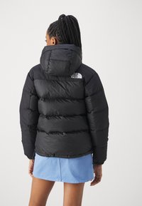 Crna puffer jakna s kapuljačom, koja ima quilted teksturu, elastične manžete i bijeli logo na leđima blizu vrata.