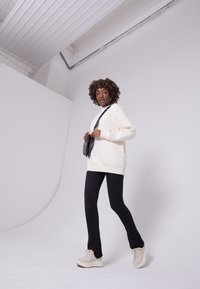 Creme oversized sweater, sorte tætsiddende leggings og lyse chunky sneakers. Modellen holder en lille sort crossbody taske. Neutral baggrund.