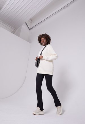 Maglione oversized color crema, leggings neri aderenti e sneakers chunky di colore chiaro. La modella tiene una piccola borsa a tracolla nera. Sfondo neutro.