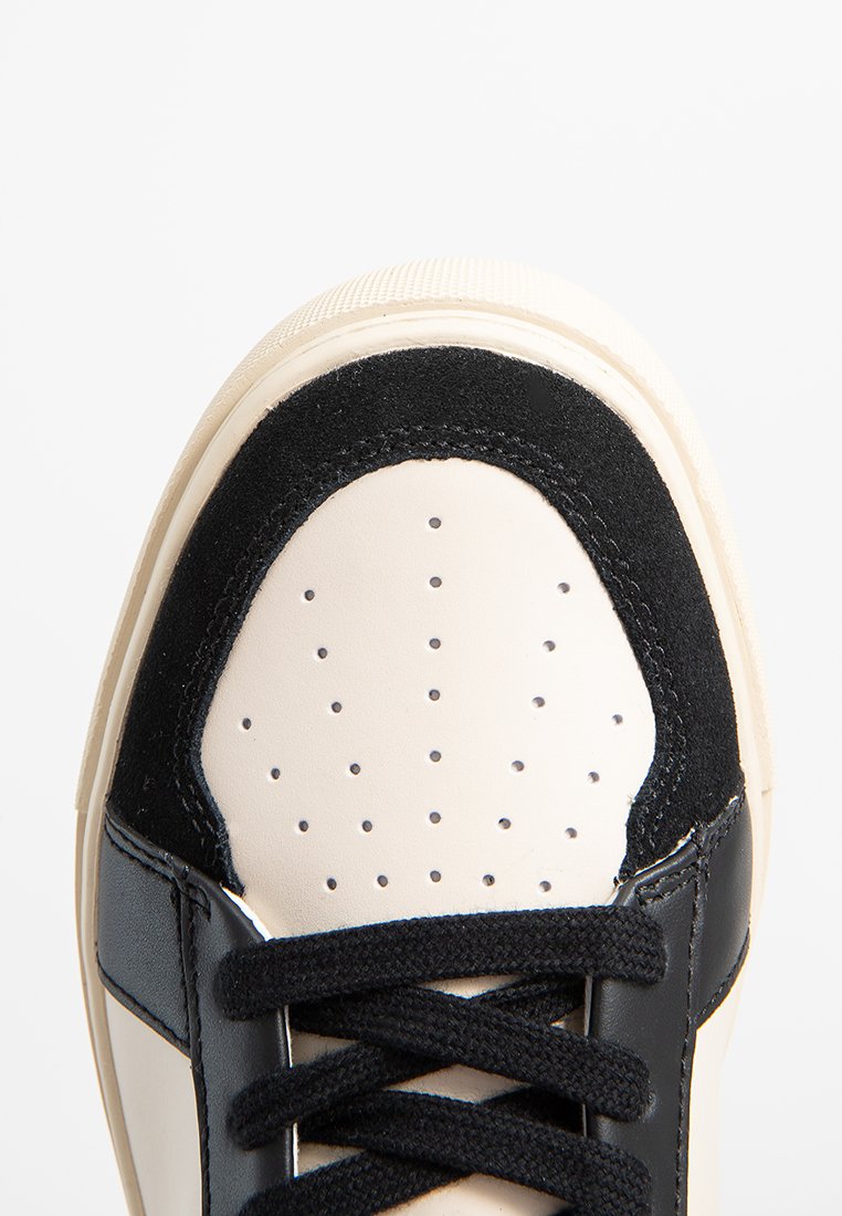 Weißer und schwarzer Sneaker mit Perforationen und schwarzen Schnürsenkeln auf einer strukturierten Gummisohle, vor einem weißen Hintergrund abgebildet.