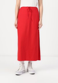 Roter, langer Maxikleid mit elastischem Bund und Kordelzug. Der Stoff ist glatt und ohne sichtbare Muster, kombiniert mit weißen Turnschuhen.