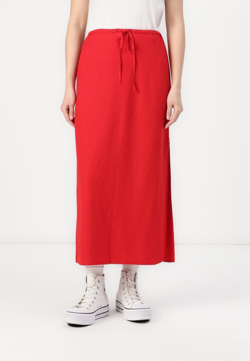 Roter, langer Maxikleid mit elastischem Bund und Kordelzug. Der Stoff ist glatt und ohne sichtbare Muster, kombiniert mit weißen Turnschuhen.