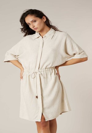 Femme posant les mains sur les hanches, portant une robe beige à manches courtes, boutonnée, avec un cordon à la taille et un col.