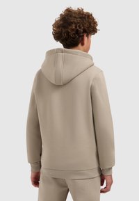 Persoon met krullend haar, gekleed in een beige hoodie en bijpassende beige broek, staand met de rug naar de camera tegen een effen achtergrond.
