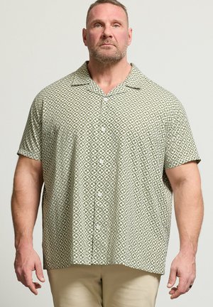Mand med kort hår og skæg iført kortærmet olivengrøn mønstret skjorte med knapper og beige bukser, stående mod en ensfarvet baggrund.