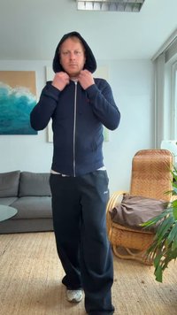 Felpa con zip di colore blu navy con cappuccio, abbinata a pantaloni neri larghi e sneakers bianche. Sfondo con opere d'arte spaziali, ambientazione interna con piante.