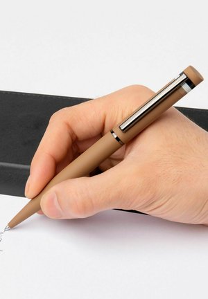 Bruine pen met een gladde, matte afwerking, metalen clip en een puntige punt, gehouden boven wit papier nabij een zwarte ondergrond.