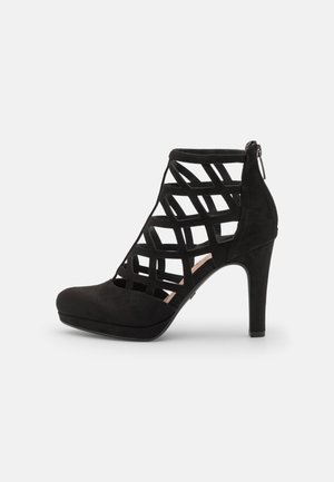 Sort ankelstøvle med høje hæle, geometrisk udskåret burdesign og lynlås bagpå, præsenteret på en enkel hvid baggrund.